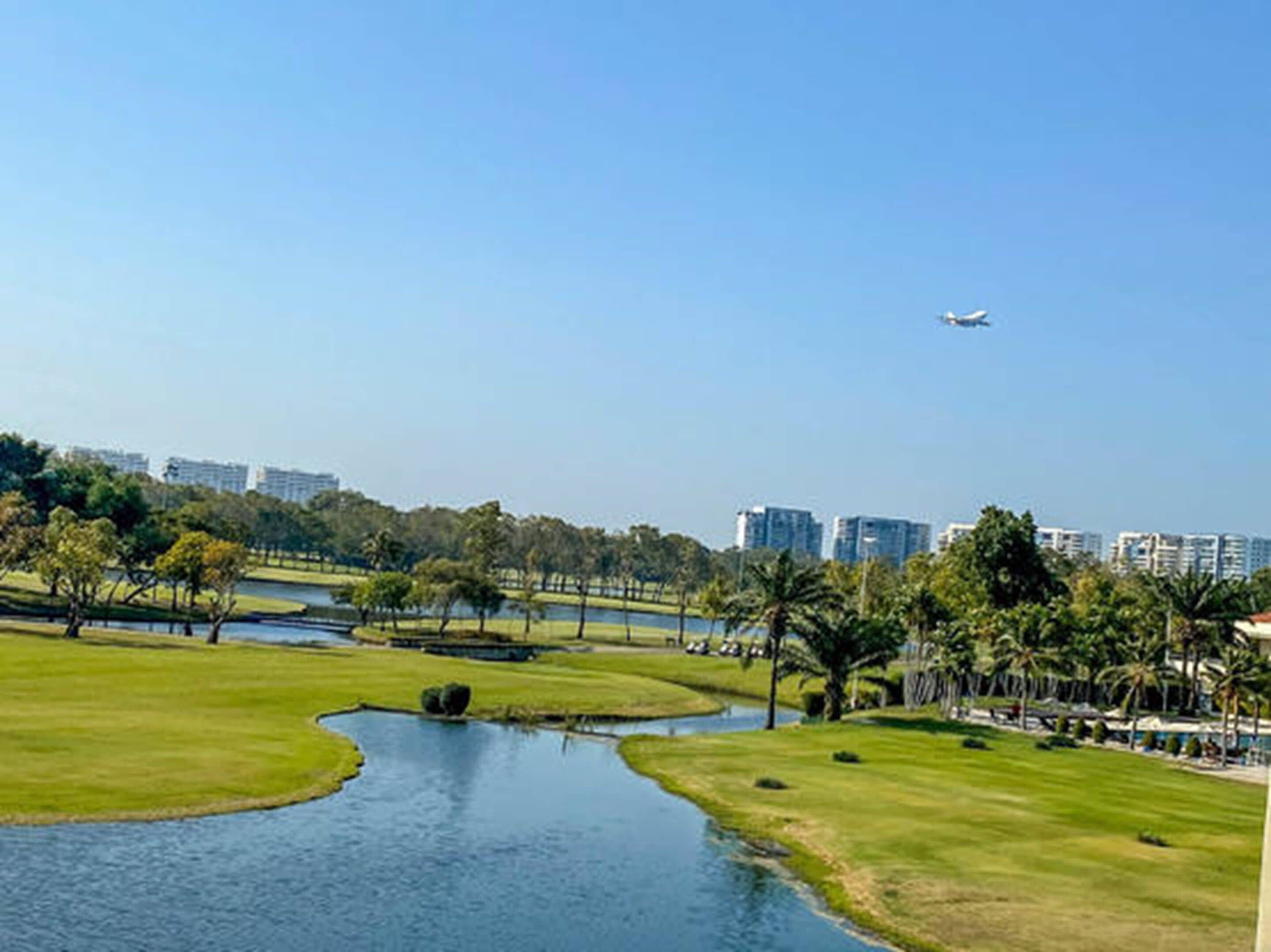 Un parcours de golf avec un plan d’eau, des palmiers et un avion qui vole au loin.