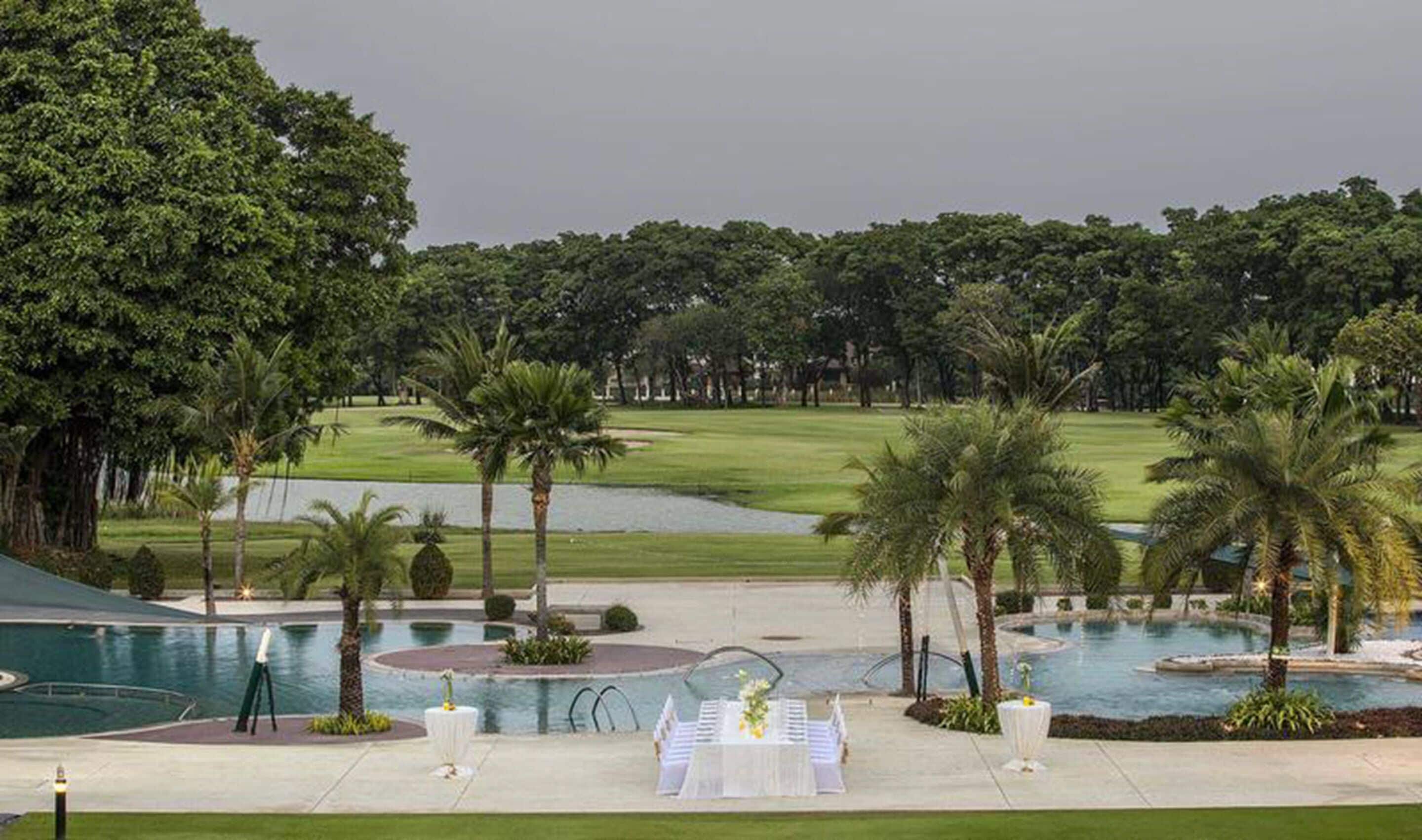 Uma área de piscina com mesa montada para um casamento, rodeada por palmeiras e um campo de golfe ao fundo.