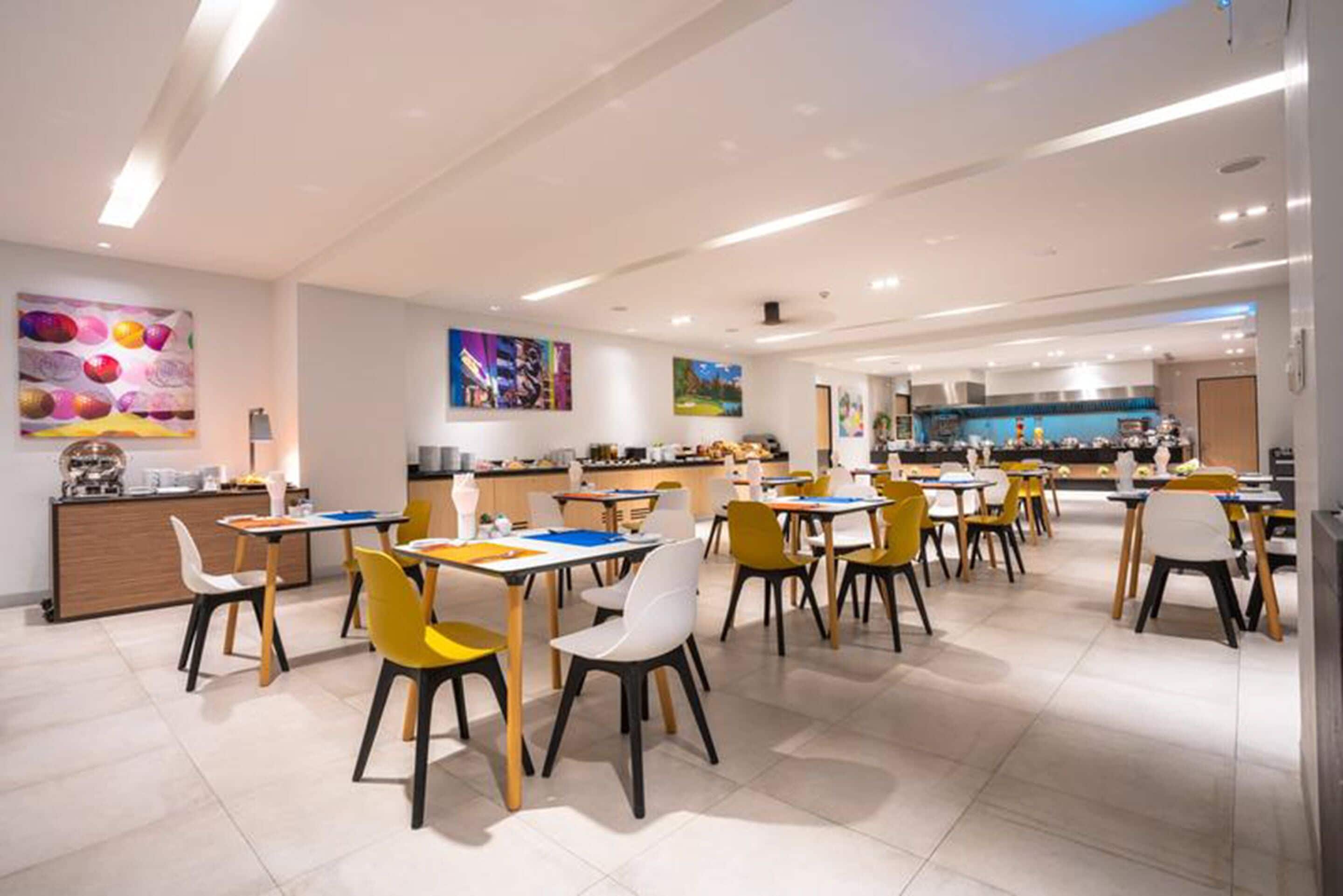 A modern dining area with multiple tables and chairs, a kitchen in the background, and artwork on the walls.