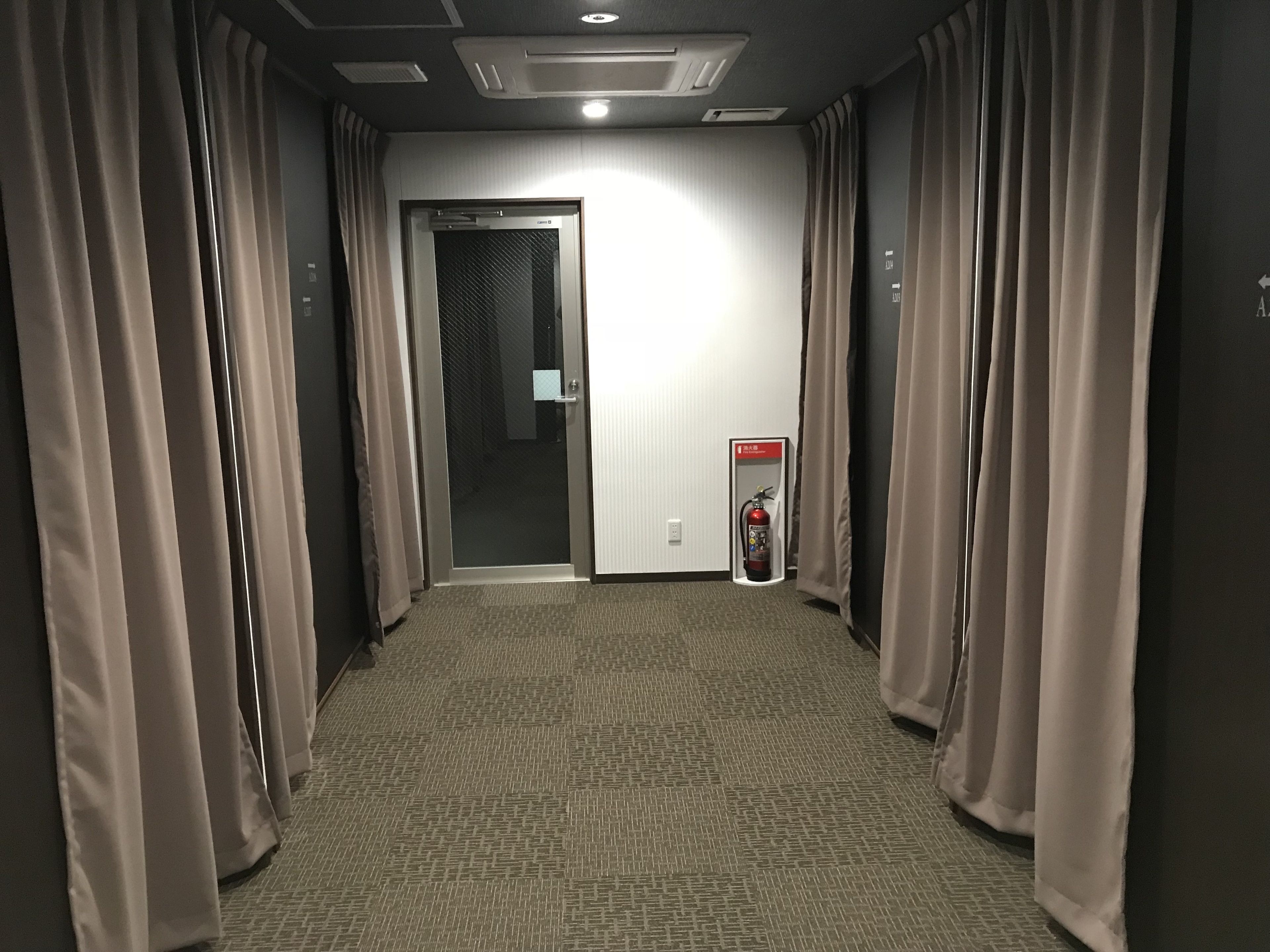 A hotel hallway with a fire extinguisher mounted on the wall, a door at the end, and curtains on both sides.