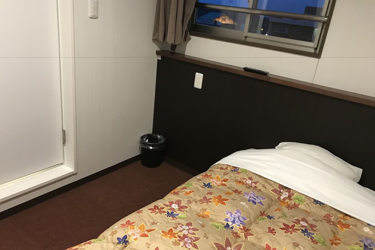 Una habitación de hotel con una cama, una ventana, una puerta y un cubo de basura.