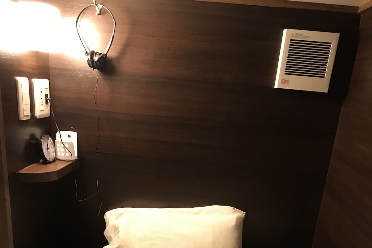 Un dormitorio con cabecera de madera, almohada blanca, colcha estampada, mesita de noche con un reloj y un calentador de pared.