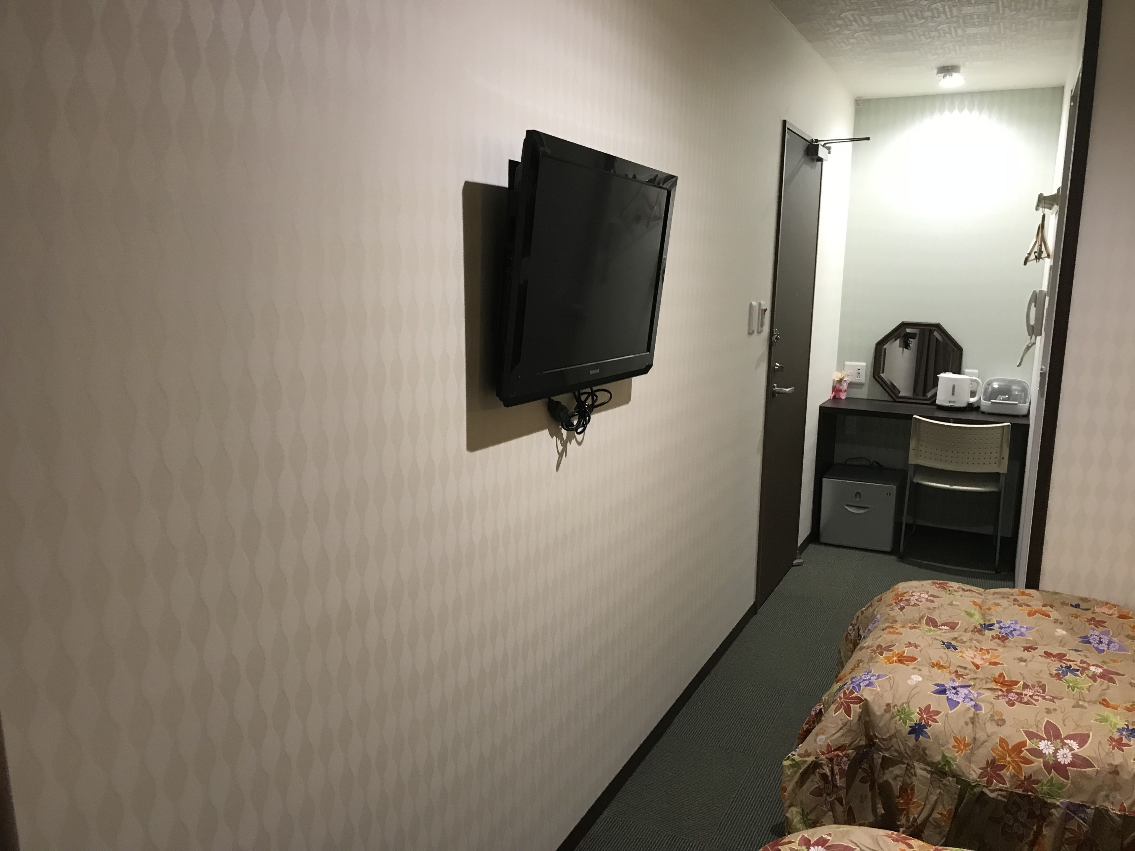 A hotel room with a television mounted on the wall, a bed with a floral bedspread, a small desk with a microwave, and a chair.