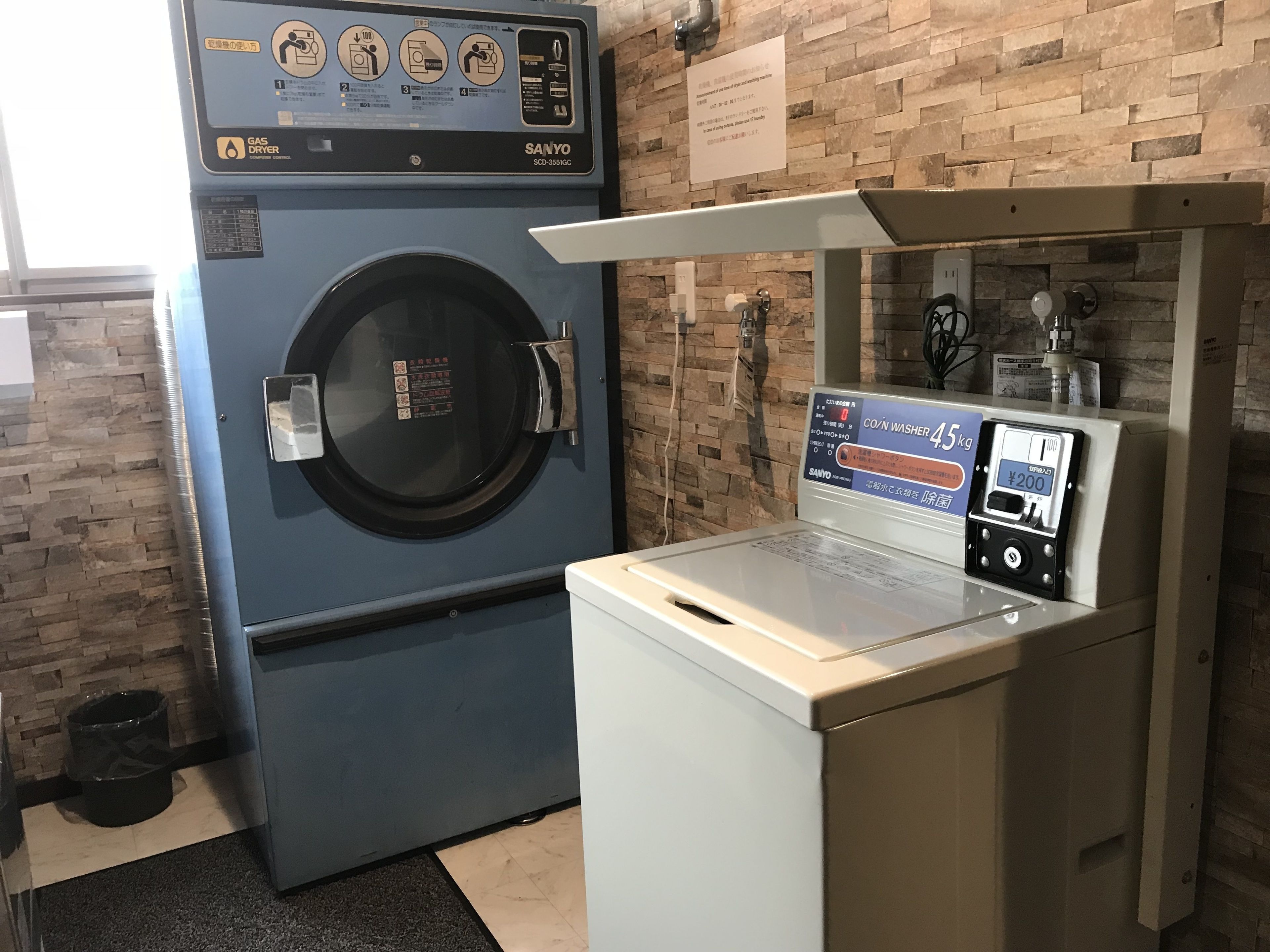A laundry facility with a blue washer and dryer, a CO/N washer, and a small trash bin.