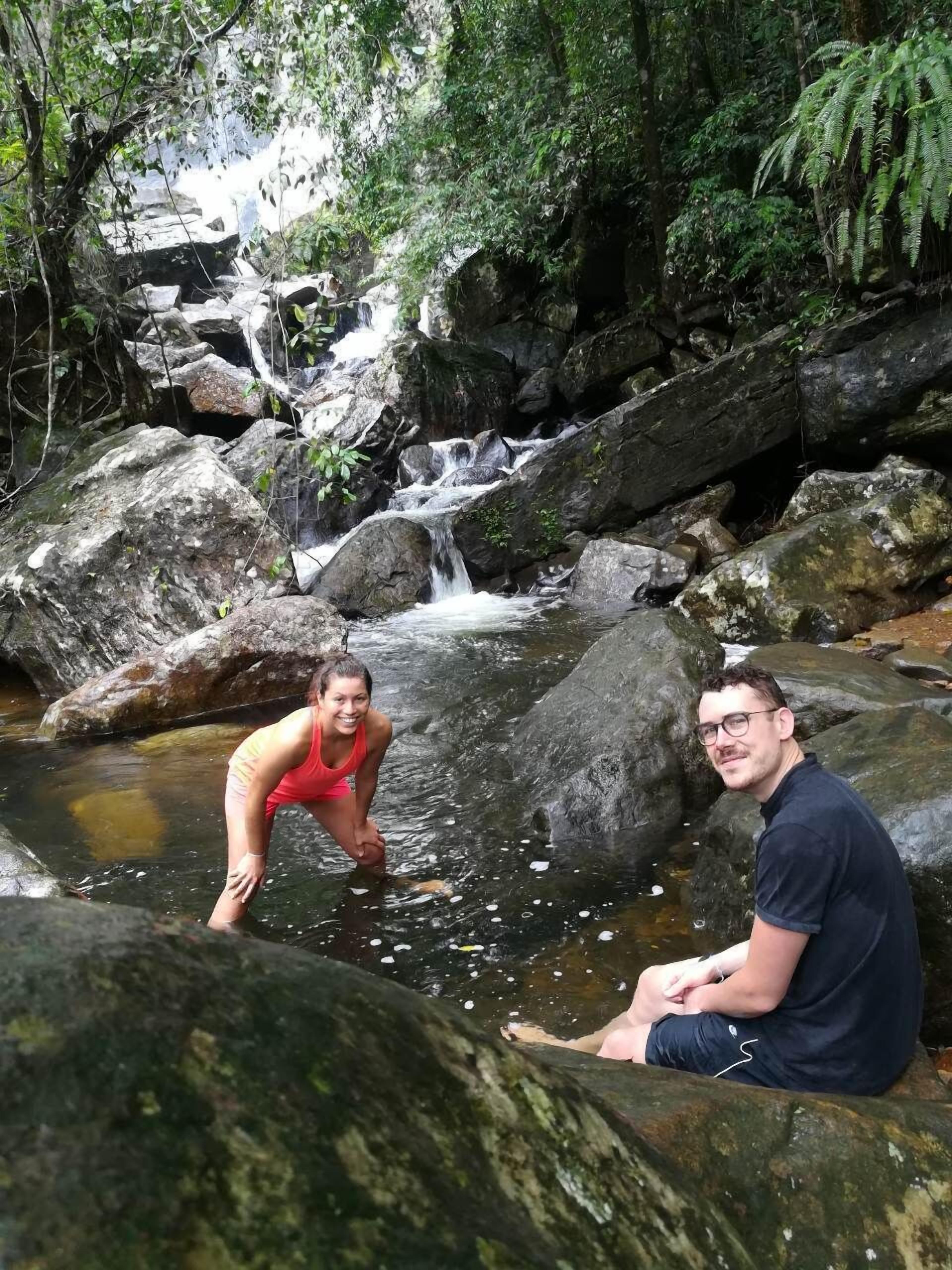Dos personas sentadas sobre rocas junto a un arroyo en un área boscosa, con una cascada al fondo.