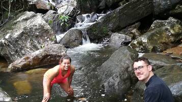 Dos personas sentadas sobre rocas junto a un arroyo en un área boscosa, con una cascada al fondo.
