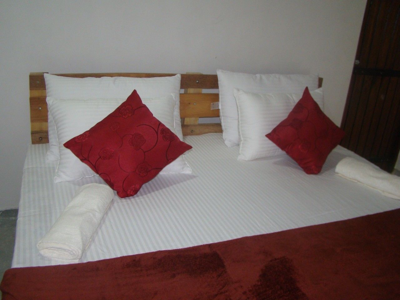 Una cama con ropa de cama blanca y dos almohadas rojas, una toalla blanca enrollada y una manta roja.