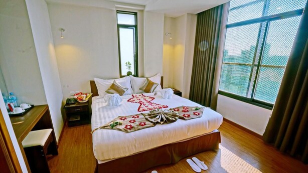 A hotel room with a large bed, a nightstand, a bedside table with a lamp, a window with a view, and a pair of slippers on the floor. - Sanctuary Hotel (Mandalay)