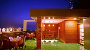 A rooftop restaurant with tables and chairs, a bar area, and a view of the city at night. - Sanctuary Hotel (Mandalay)