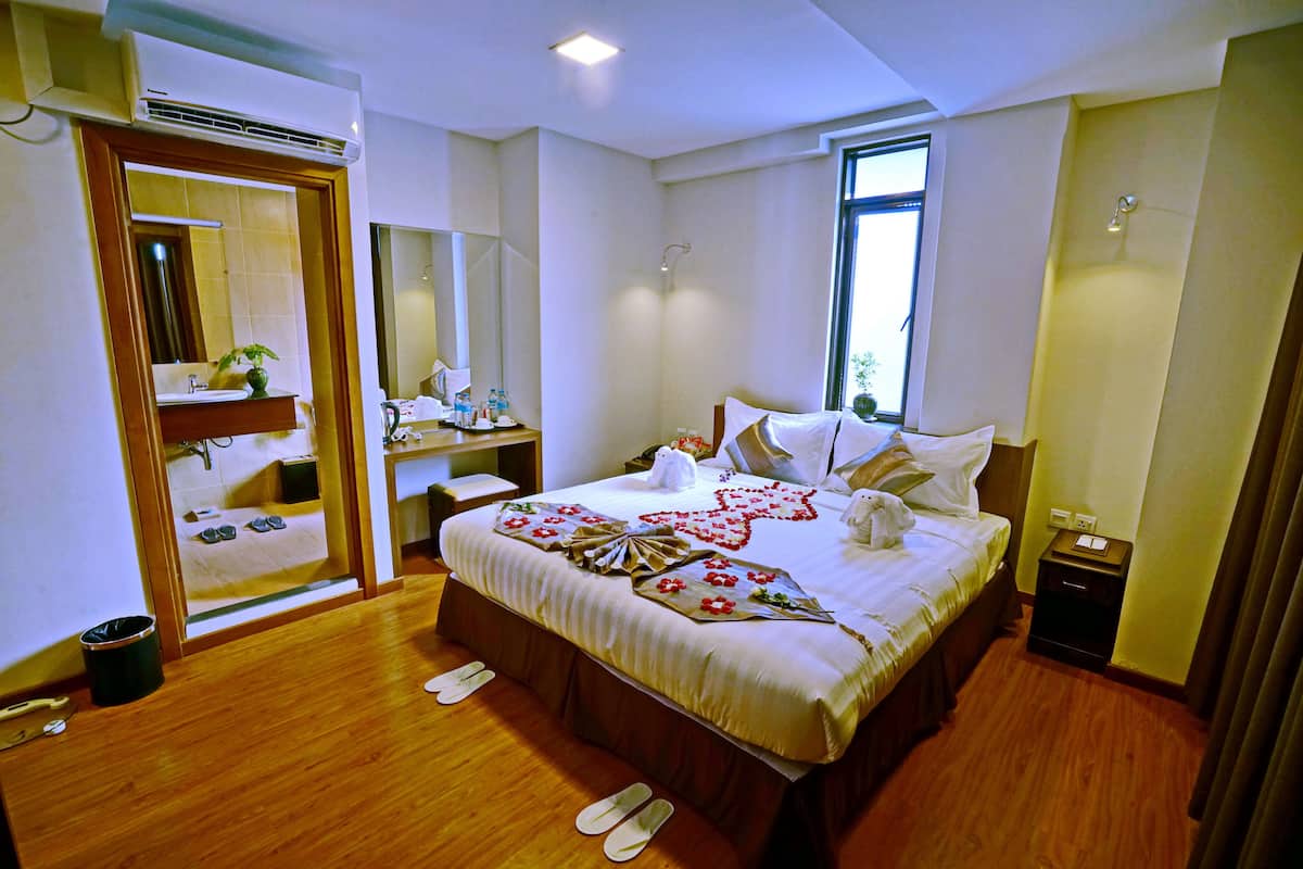 A hotel room with a large bed, a bedside table, a mirror, a window, and a wooden floor.
