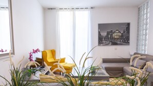 A modern living room with a yellow armchair, a grey sofa, and a glass coffee table. There are potted plants in the foreground and a cityscape painting on the wall.