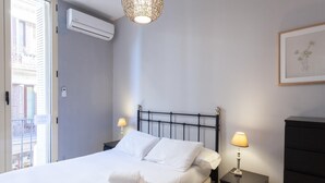 A neatly made bed with white linens and two pillows, a black metal headboard, bedside tables with lamps, a black dresser, and an air conditioning unit on the wall.