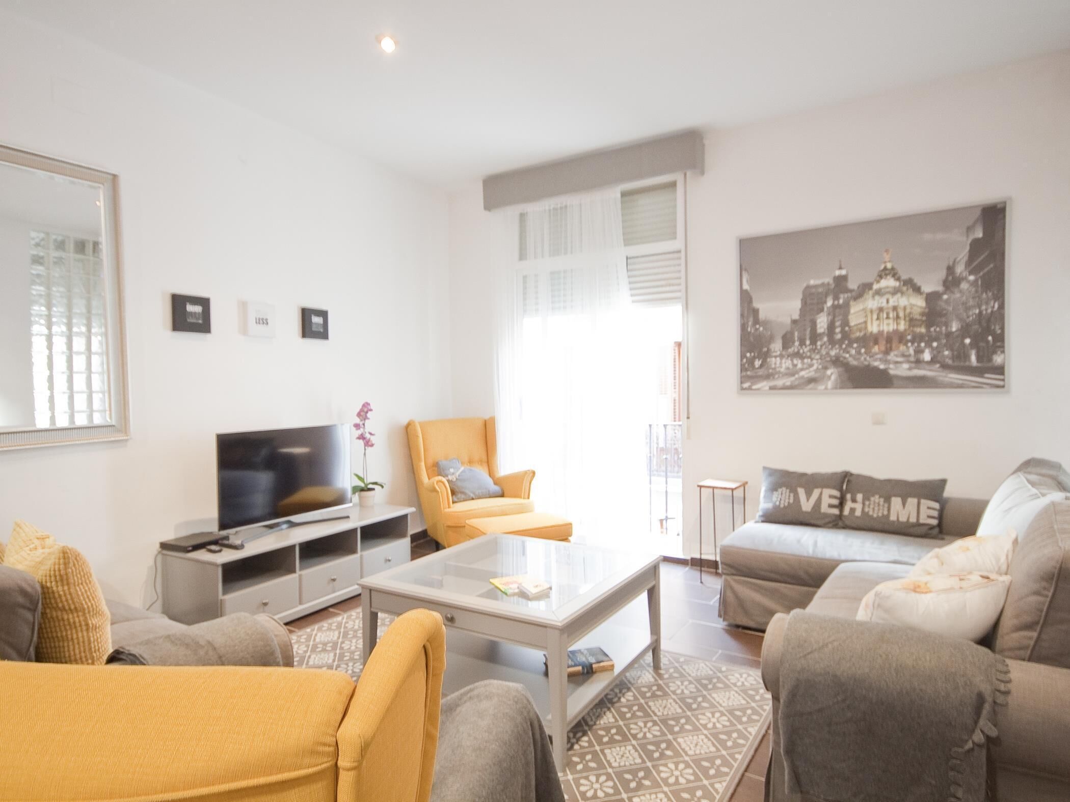 Una sala de estar moderna con sofá, sillón y una mesita de centro. Hay un televisor, una pequeña mesa con una planta y un cuadro de un paisaje urbano en la pared.