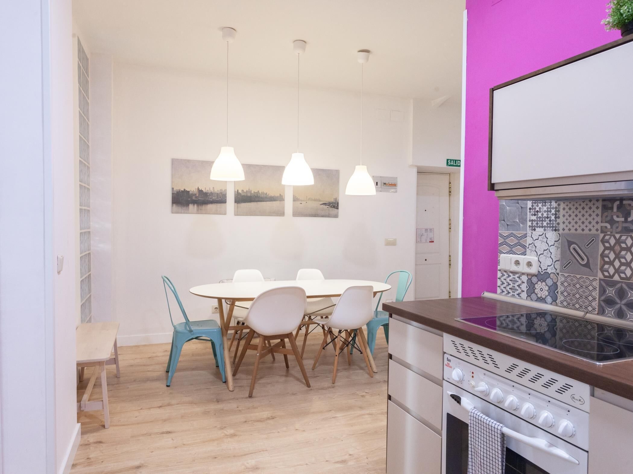 Une cuisine moderne dotée d’un four encastré, d’un coin repas avec une table et des chaises blanches, et d’un mur d’accent rose.