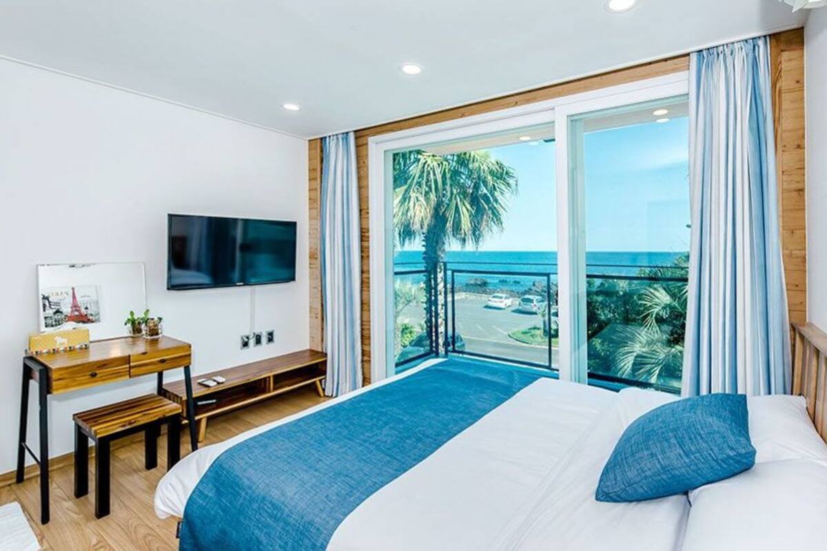 A hotel room with a bed, a desk, a TV, and a balcony with a view of the ocean.