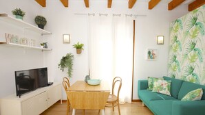 A modern living room with a wooden dining table, chairs, a sofa, and a TV on a cabinet. There are plants on shelves and a window with curtains. - Bright Studio in the heart of Palma with Balcony, Wifi, AC, Great for Couples (Palma)