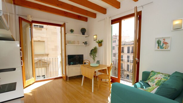 A living room with a green sofa, wooden flooring, a dining table with chairs, a TV, and a balcony with a view of buildings. - Bright Studio in the heart of Palma with Balcony, Wifi, AC, Great for Couples (Palma)