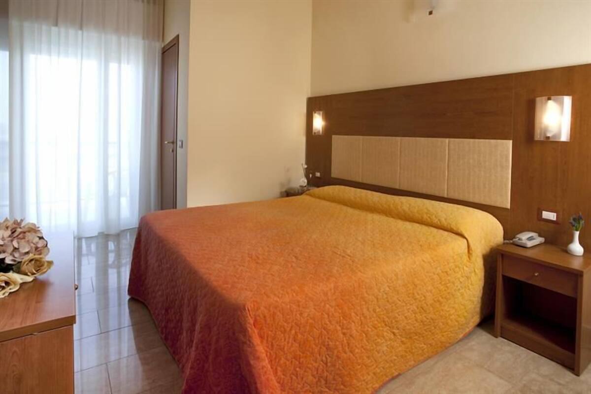 A hotel room with a large bed, a bedside table, a nightstand, a wall-mounted light, and a door.