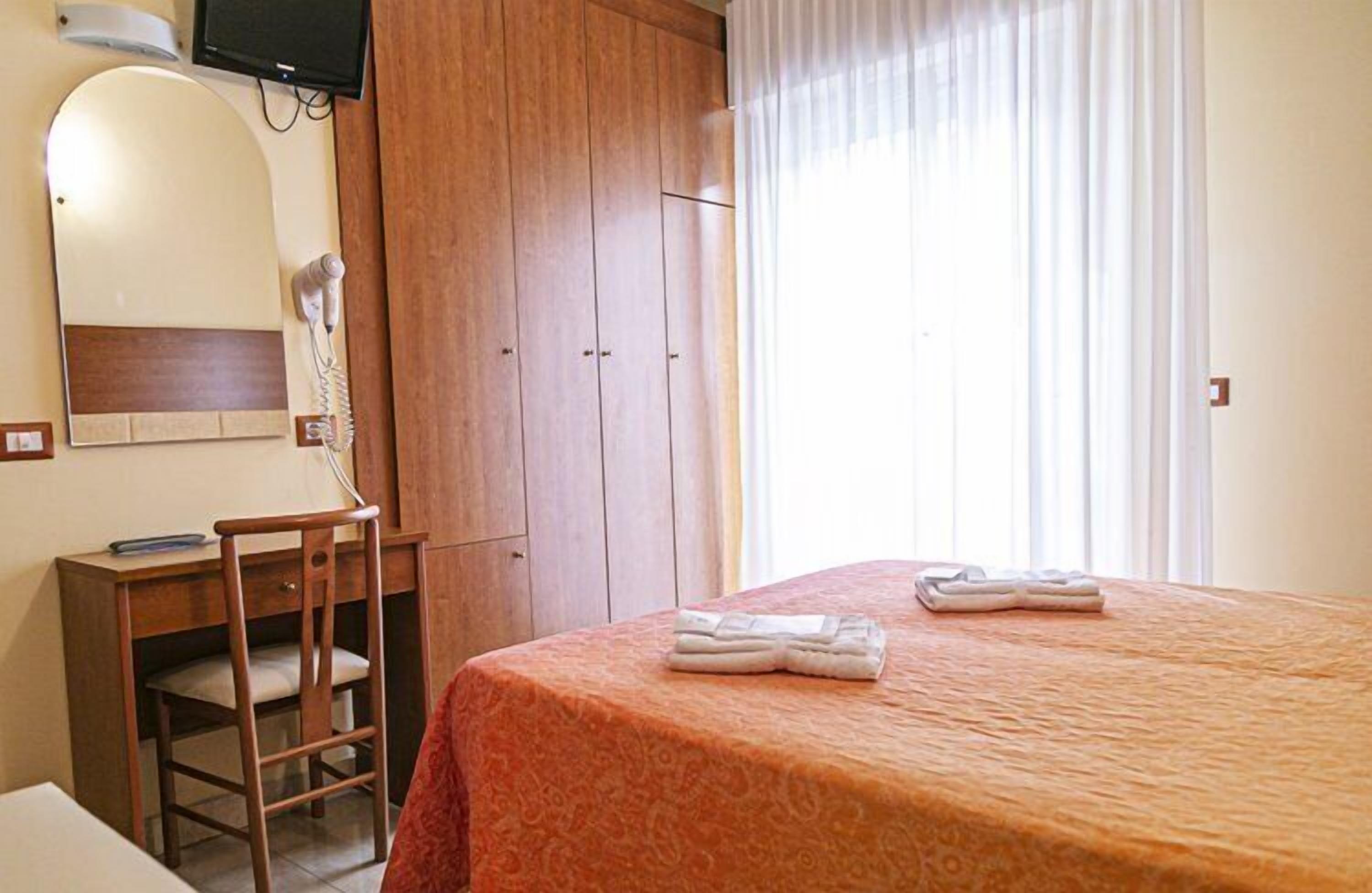 A hotel room with a bed, a desk with a chair, a mirror, a hairdryer, and a wardrobe.
