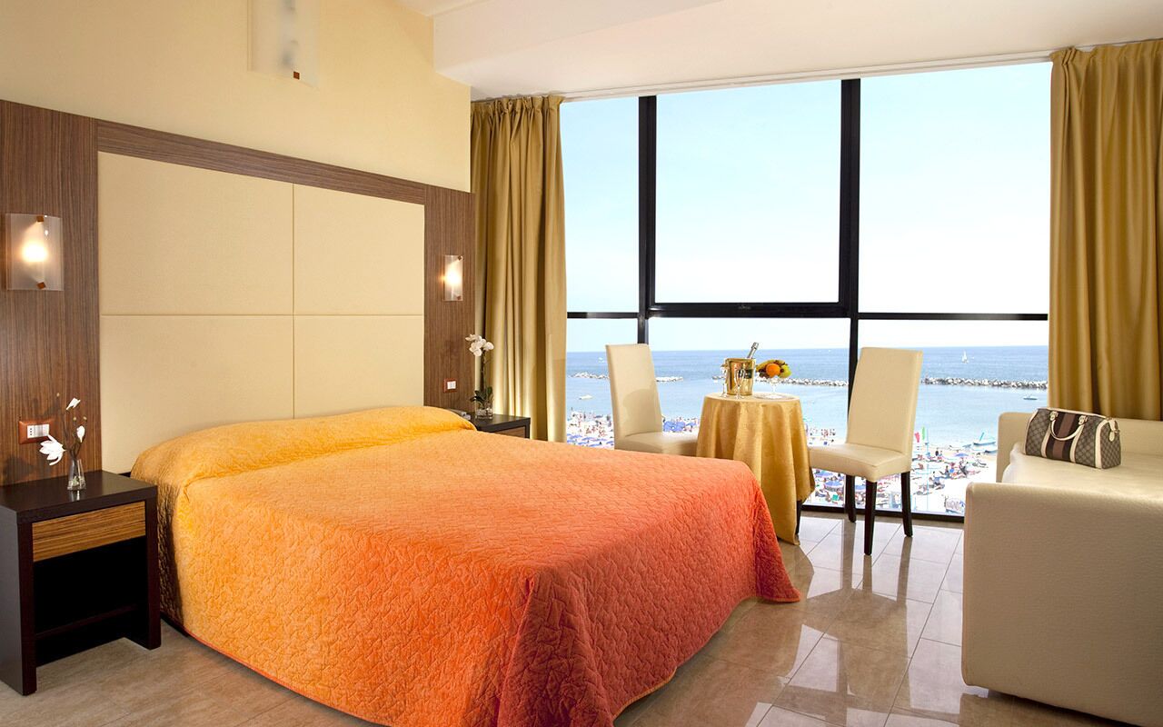 Habitación de hotel con una cama grande, zona de estar con vista al mar y una mesita con un florero.