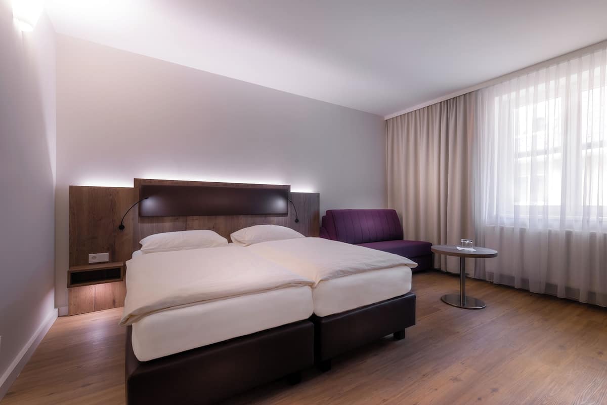 A modern hotel room with a large bed, a small round table, a purple armchair, and a large window with sheer curtains.