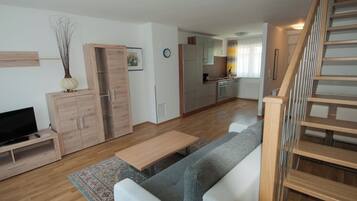 Una sala de estar moderna con un sofá, televisión, armarios de madera y una cocina al fondo.