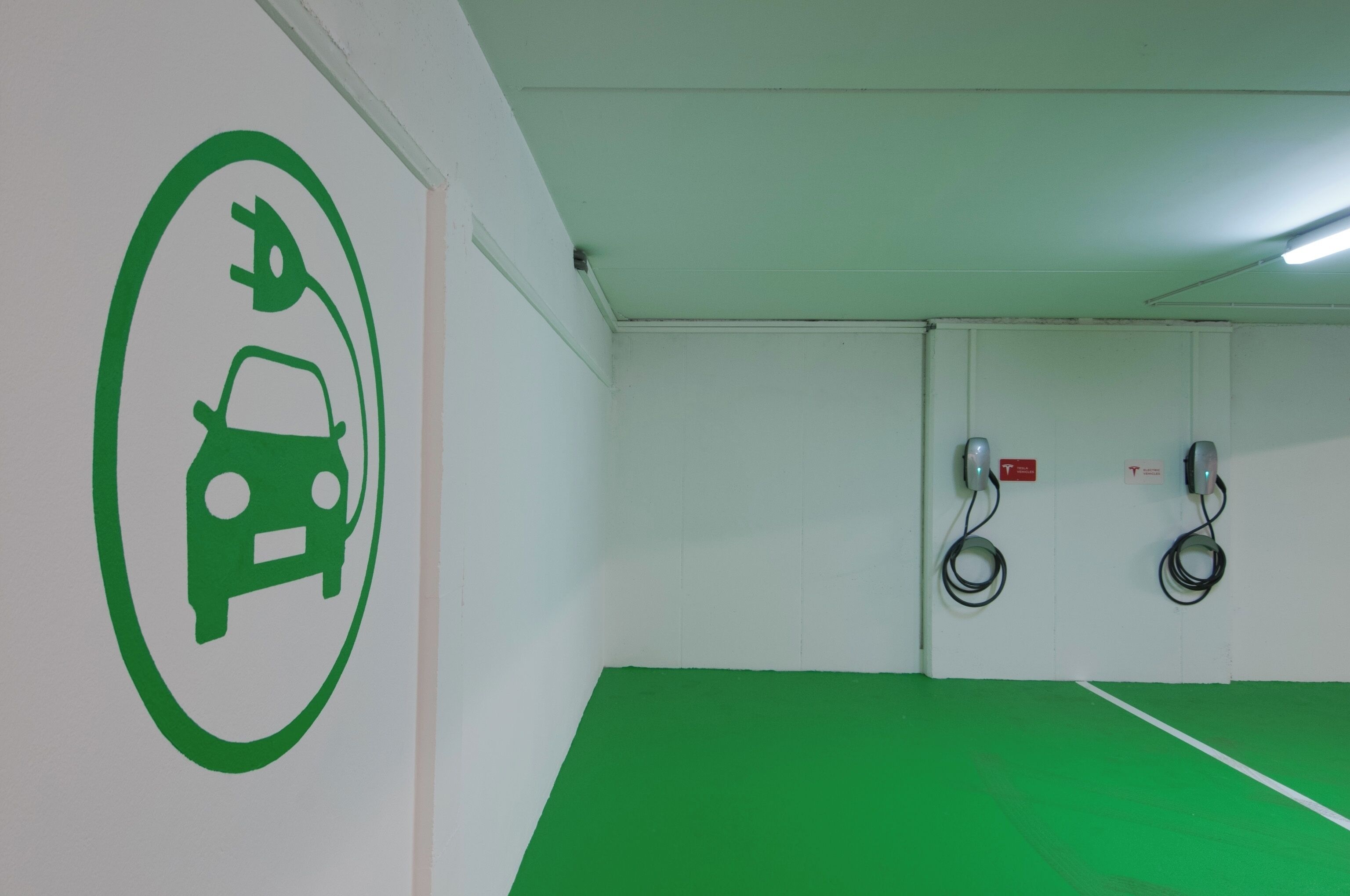 Un área de estacionamiento con suelo verde, una estación de carga para vehículos eléctricos montada en la pared e icono verde que indica la disponibilidad de carga eléctrica.