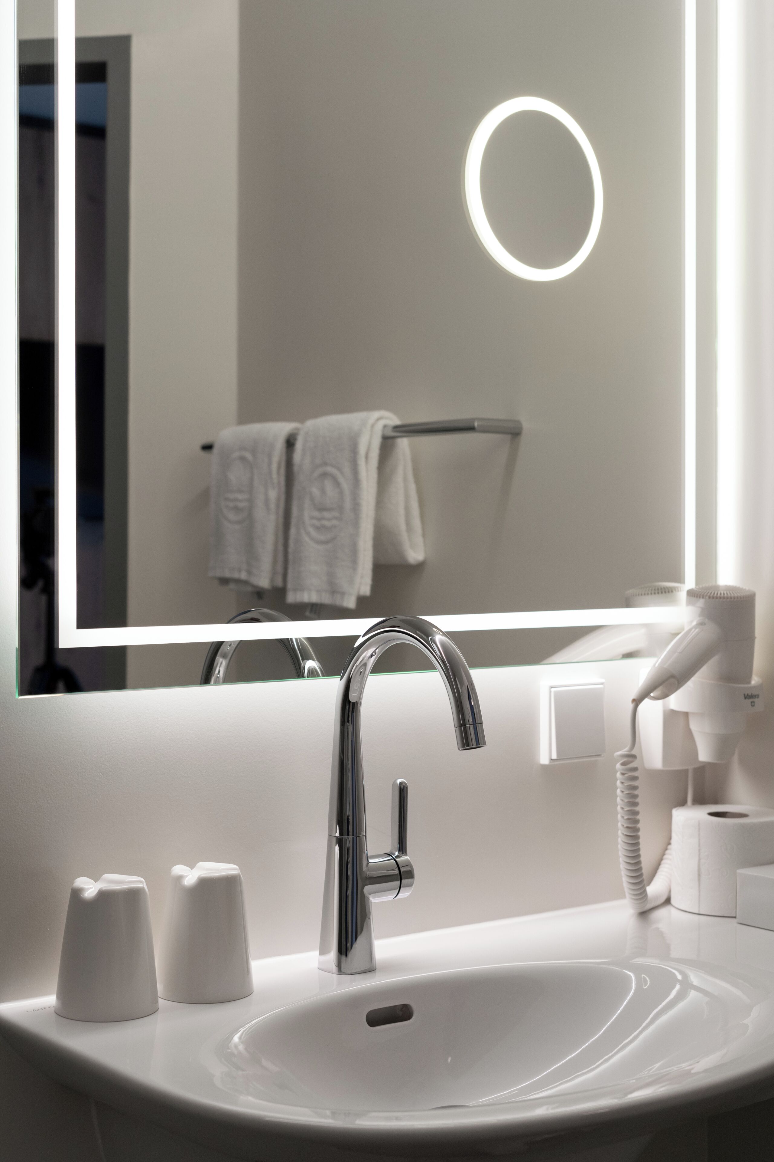 Een moderne badkamer met een wit wastafelblad, een chromen kraan en een spiegel met ronde verlichting.