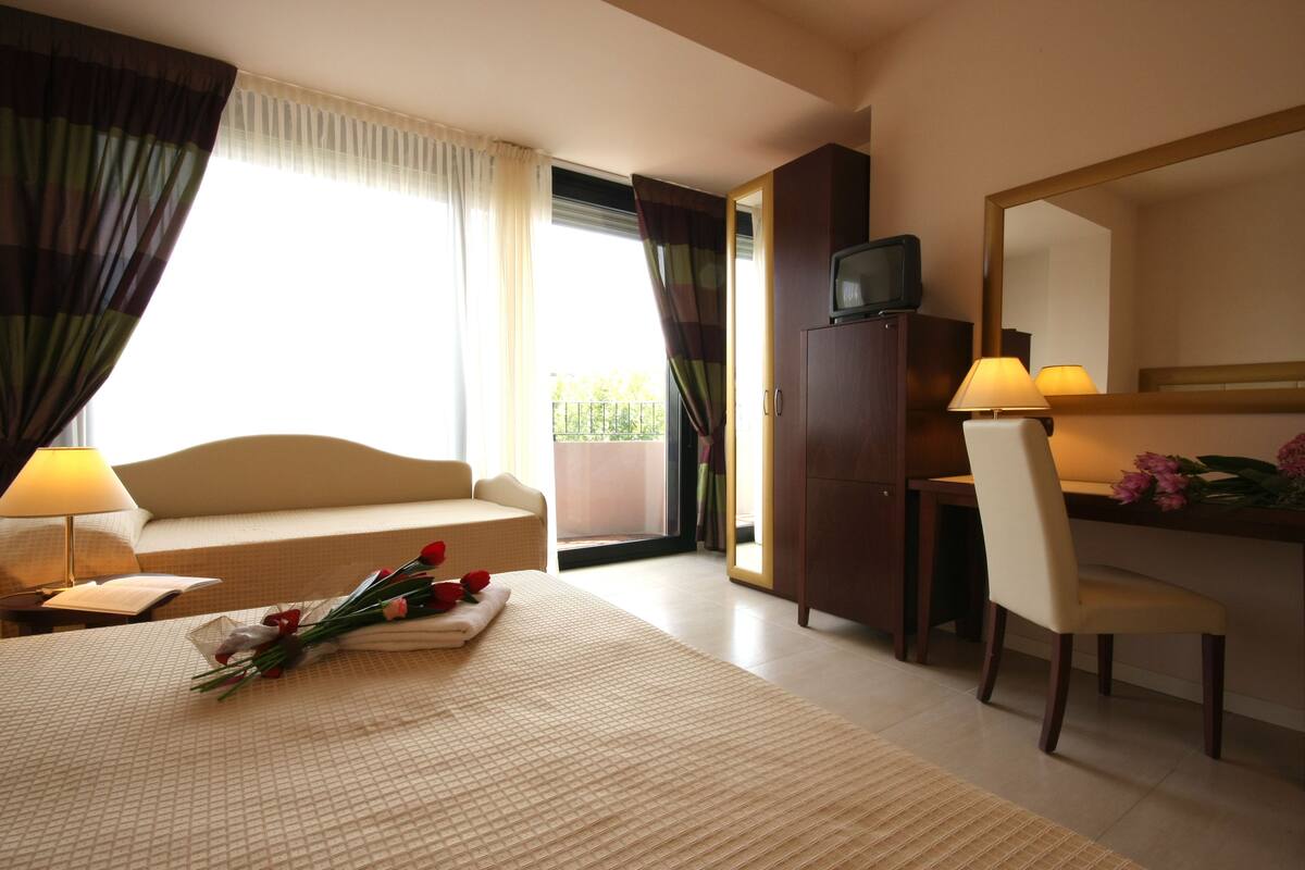 Ein Hotelzimmer mit Bett, Schreibtisch, Stuhl, Fernseher und einem Fenster mit Vorhängen.
