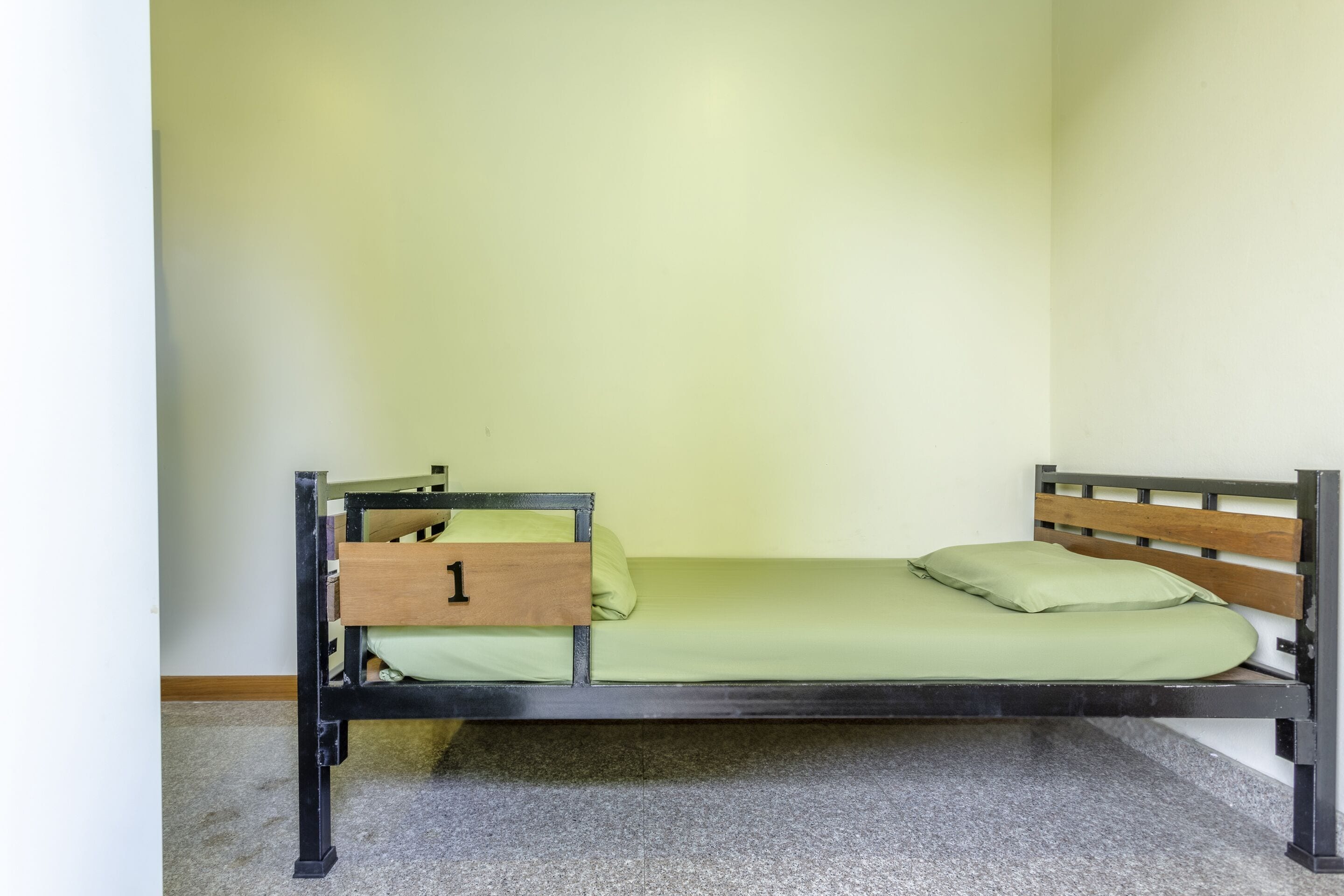 A single bed with a wooden headboard and a green mattress, a wooden nightstand with a drawer, and a carpeted floor.
