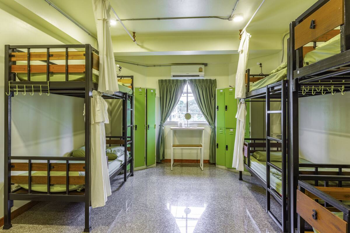 A dormitory room with bunk beds, a small desk, and a window with curtains.
