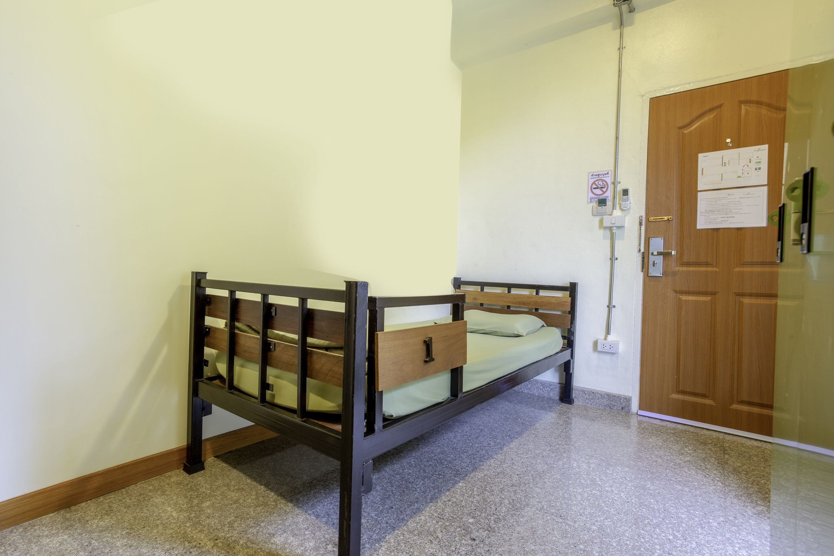 Un letto singolo con struttura in legno e cassetto estraibile, una porta con un avviso e una presa elettrica a muro.