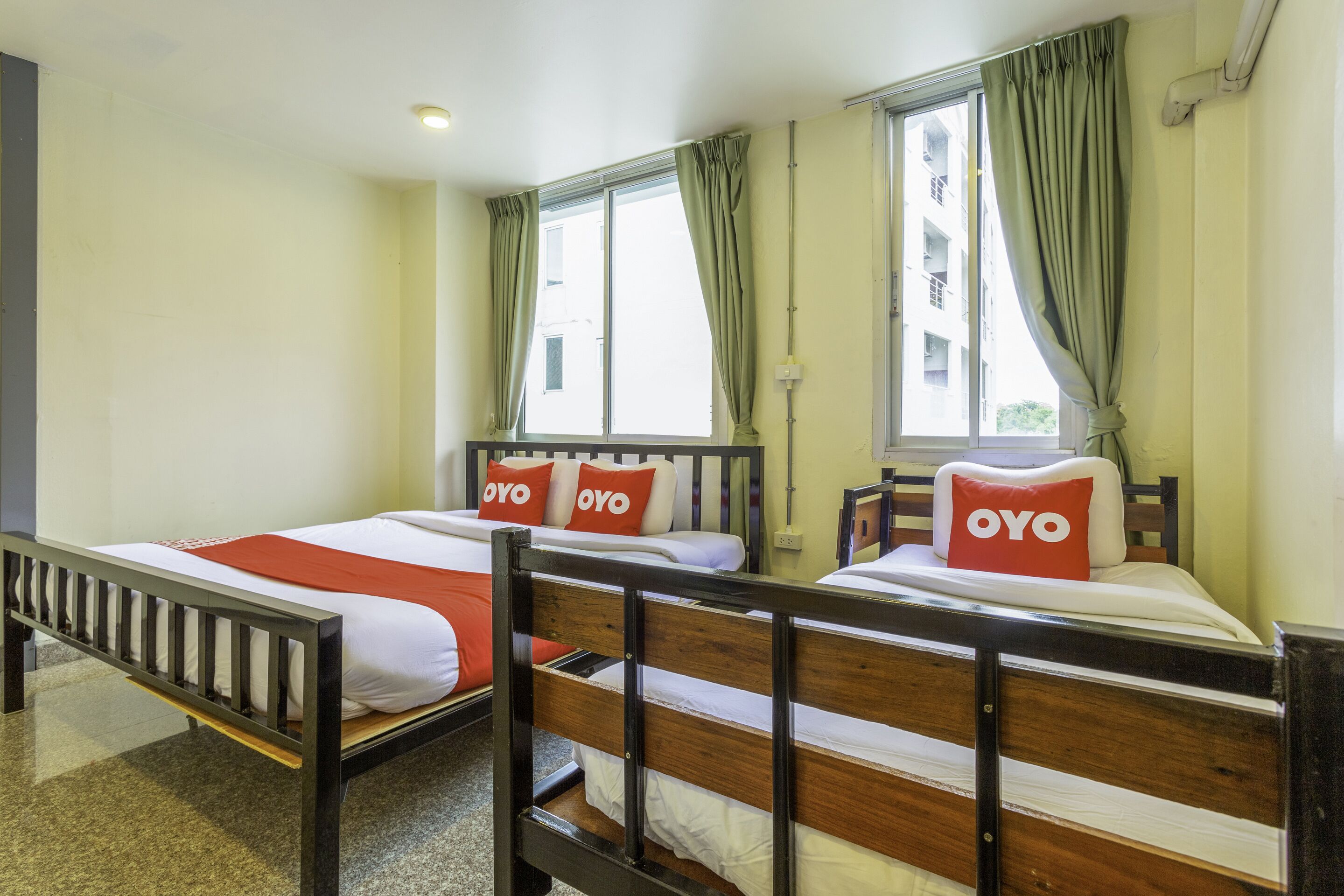 A hotel room with two beds, each with a wooden frame and white bedding. There are red pillows with the word "OYO" on them. The room has large windows with green curtains, and a view of buildings outside.