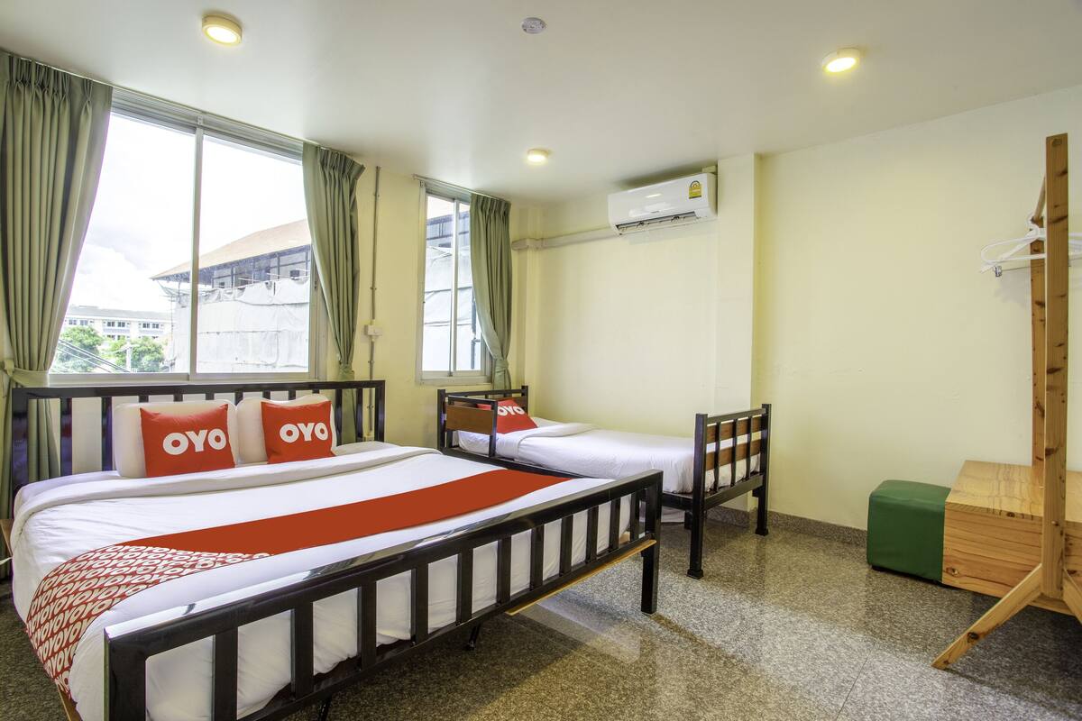 A hotel room with two single beds, a wooden chair, a green ottoman, and an air conditioning unit on the wall.