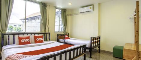 Quarto de hotel com duas camas de solteiro, uma cadeira de madeira, um banquinho verde e uma escrivaninha de madeira.