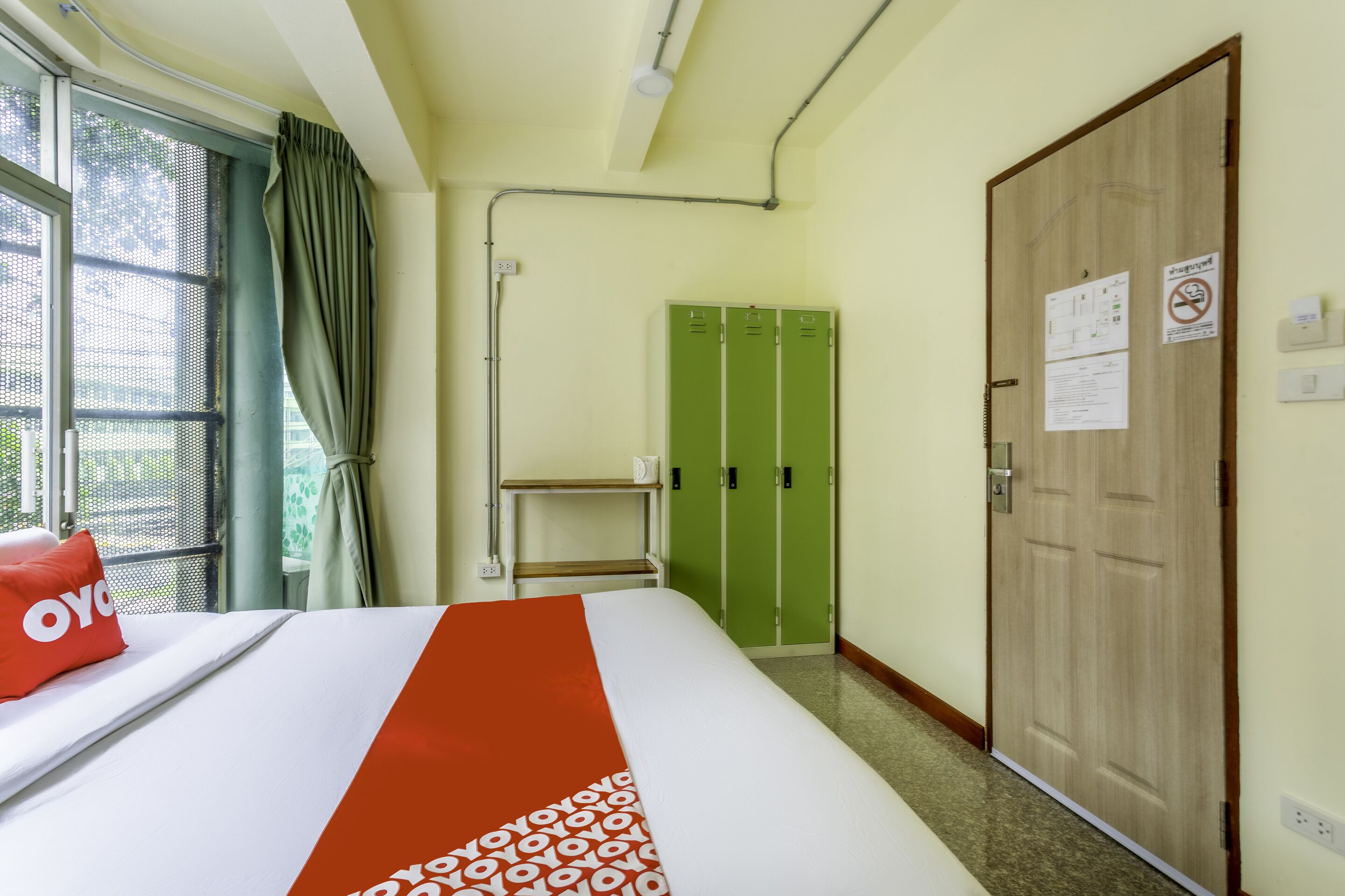 Une chambre d’hôtel avec un lit, une fenêtre avec des rideaux, une armoire verte et une porte en bois.