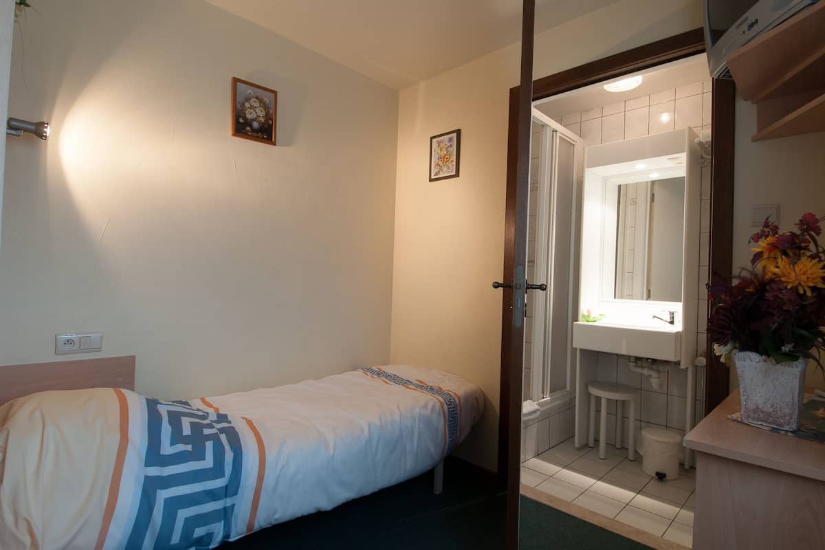Ein kleines, helles Schlafzimmer mit einem Einzelbett, ein Badezimmer mit Waschbecken und Spiegel sowie ein kleiner Hocker.