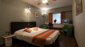 A hotel room with a bed, a desk, a chair, a television, and a ceiling fan. - Hotel Apollo (Zutendaal)