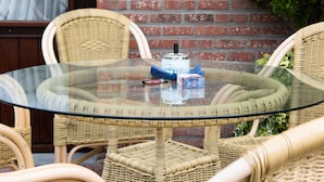 A round glass table surrounded by wicker chairs in an outdoor setting with a brick wall and greenery in the background. - Hotel Apollo (Zutendaal)