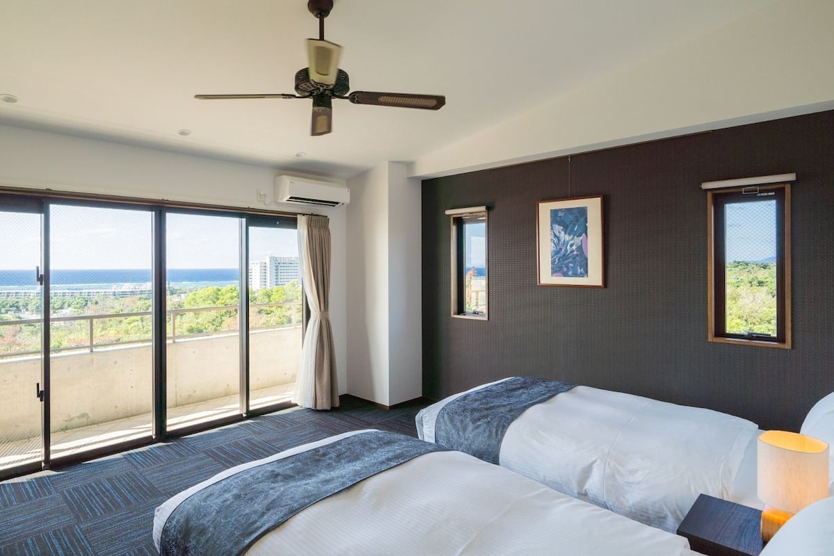 Habitación de hotel con un balcón grande, ventilador de techo y dos camas con ropa de cama azul.