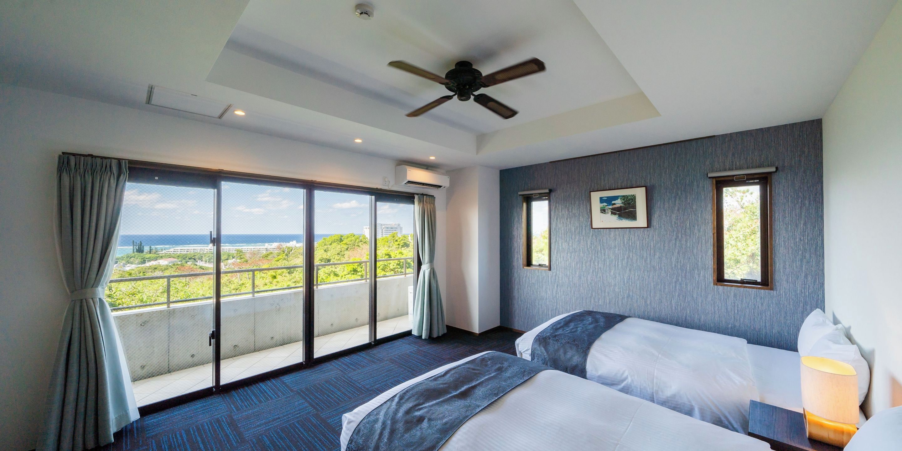 Habitación de hotel con dos camas, ventilador de techo, ventanas amplias con cortinas y vista al mar.