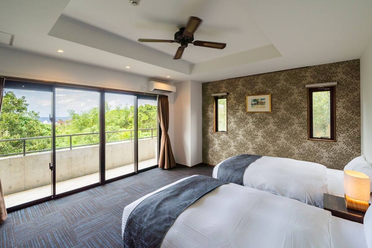 Habitación de hotel con dos camas, un ventilador de techo, ventanas grandes con cortinas, un cuadro en la pared y vistas a la vegetación exterior.