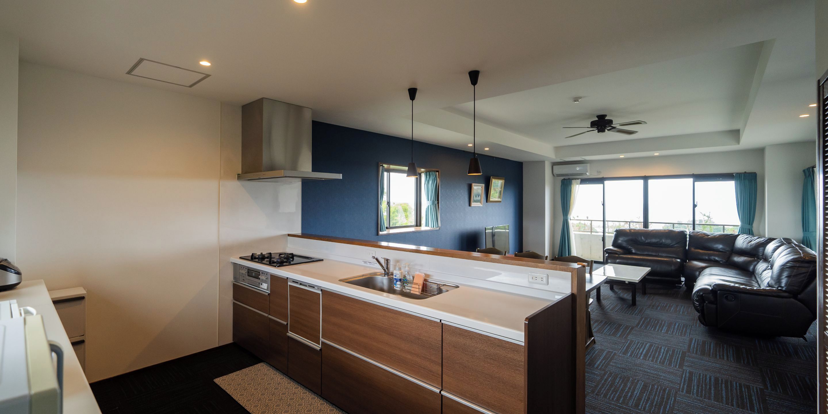 A modern kitchen with a central island, a dining area with a black leather sofa, and a view of the outdoors through large windows.