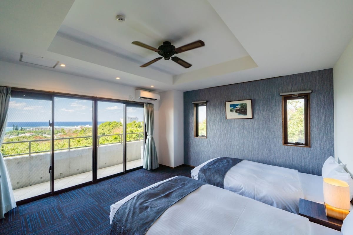 Habitación de hotel con dos camas, ventilador de techo, ventanas grandes con cortinas y vista al océano.