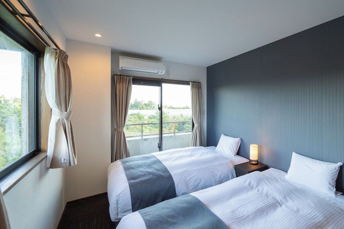 Quarto de hotel com duas camas, uma janela com cortinas, um ar condicionado e uma mesa de cabeceira com abajur.