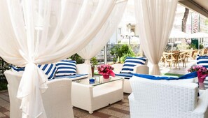 A sunlit outdoor seating area with white wicker furniture, blue-striped cushions, and a glass table. - Hotel Capinera (Rimini)