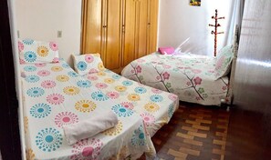 A room with two beds, each with colorful floral bedding. There is a wooden wardrobe and a coat rack.