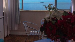 A room with a large window offering a view of the sea, a metal chair, a glass table, and a vase with flowers. - Aconchego Frente Mar (Rio de Janeiro)