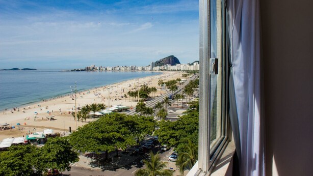 A beach view from a window overlooking a sandy beach with palm trees, a road, and buildings in the distance. - Aconchego Frente Mar (Rio de Janeiro)