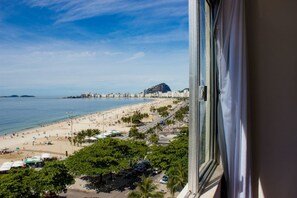 A beach view from a window overlooking a sandy beach with palm trees, a road, and buildings in the distance. - Aconchego Frente Mar (Rio de Janeiro)